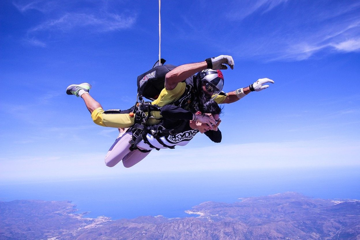 Two skydivers in freefall over a coastline under a clear blue sky.