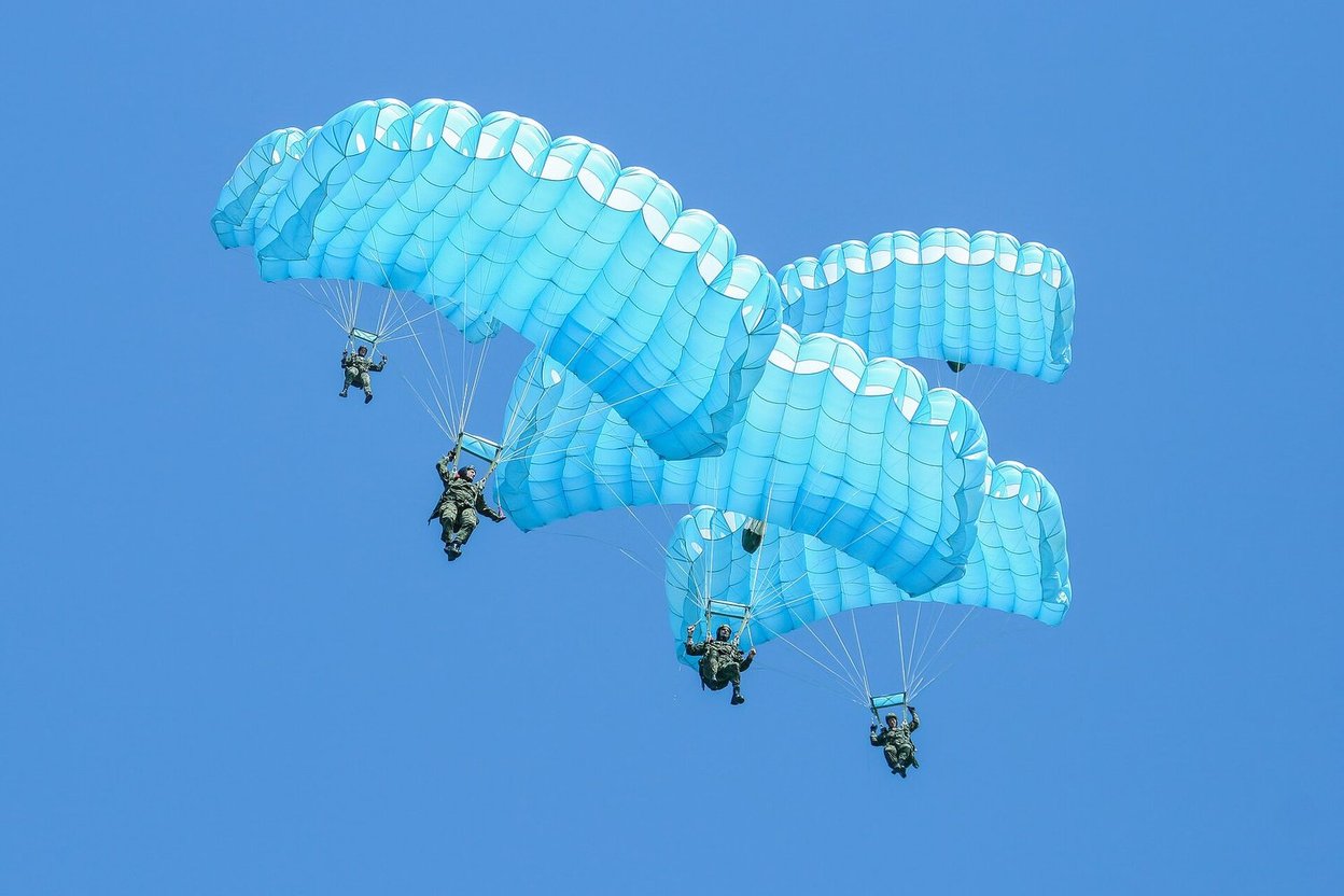 Three skydivers under light blue parachutes against a clear blue sky