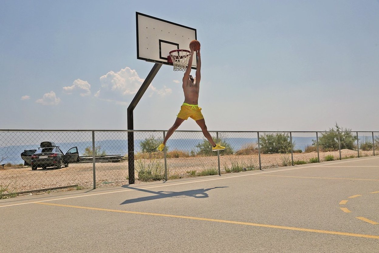 A player leaping to dunk a basketball on an outdoor court, with the coast in the background.