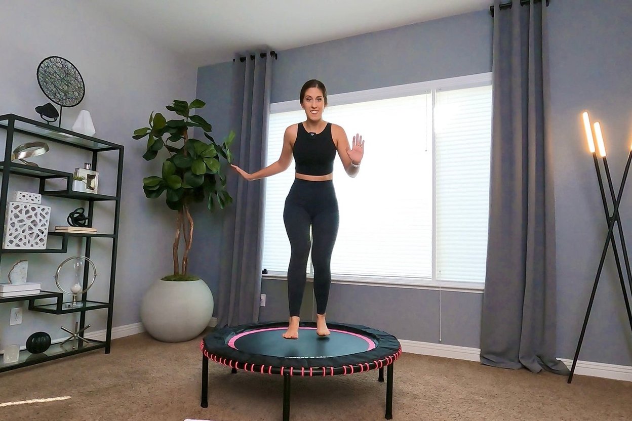 Adult doing a low health bounce on a round bungee-cord mini-trampoline in a living room, both hands on a padded stability bar, knees slightly bent
