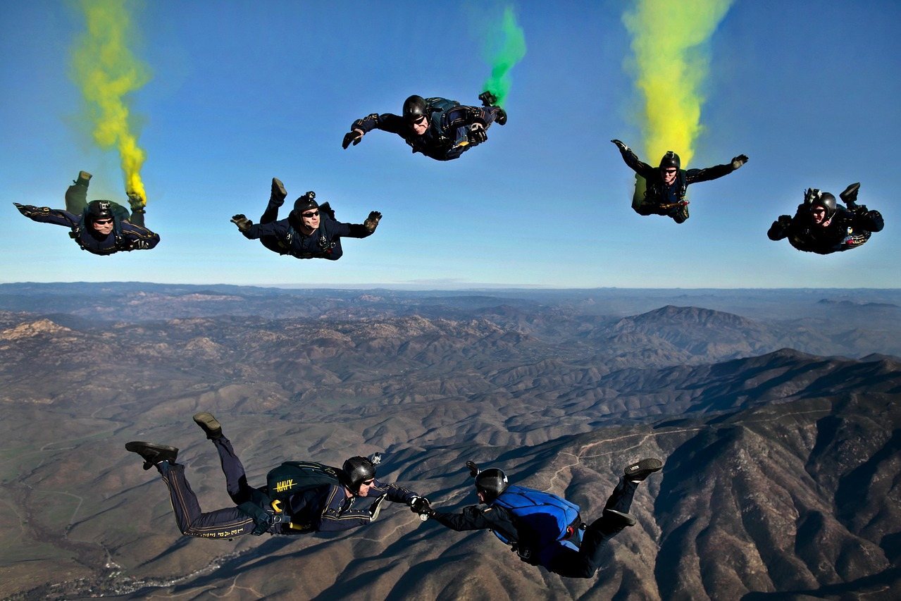 Six skydivers in freefall with colored smoke trails over mountainous terrain