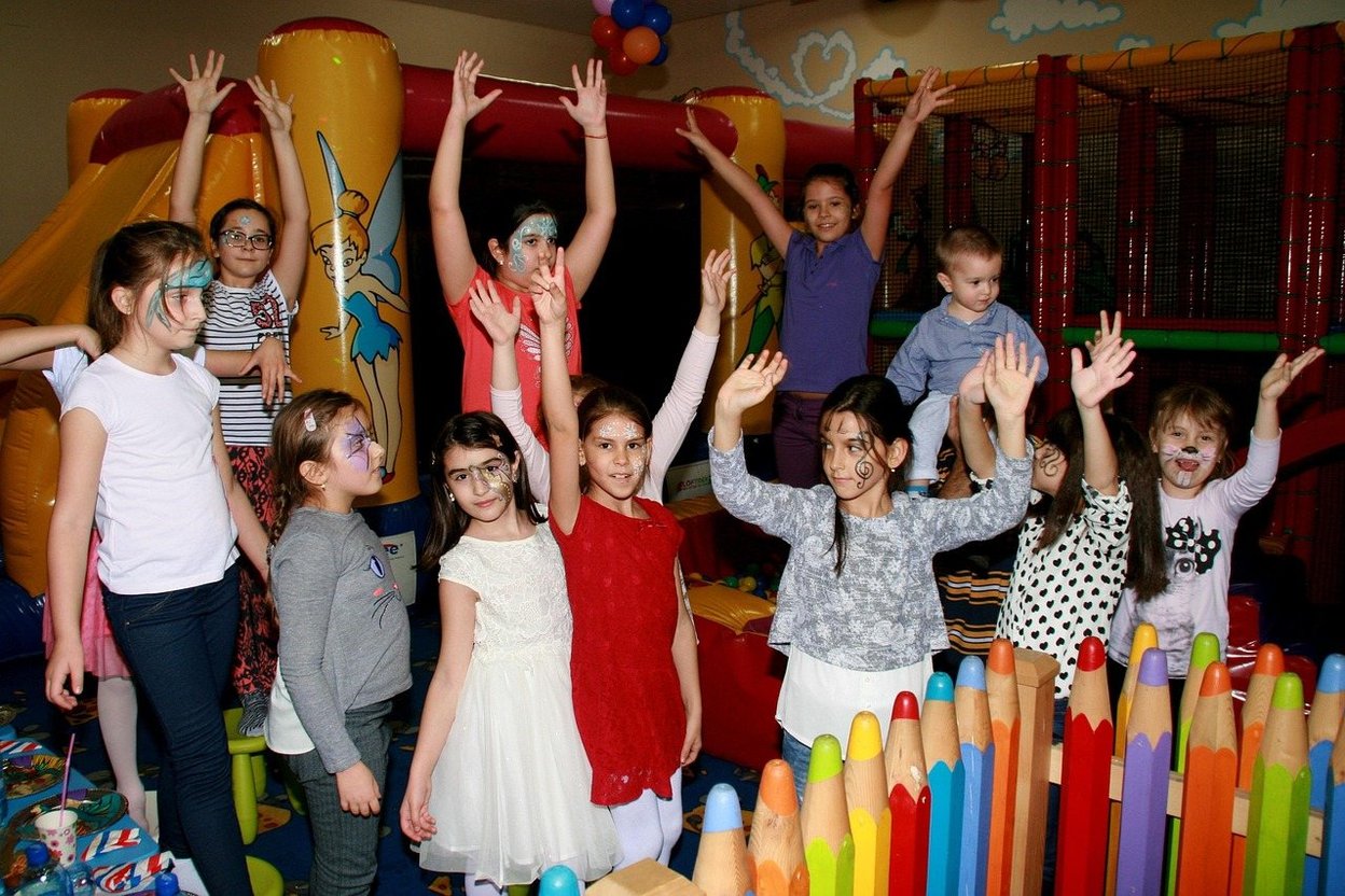 Children with painted faces raise hands in a colorful indoor play area.