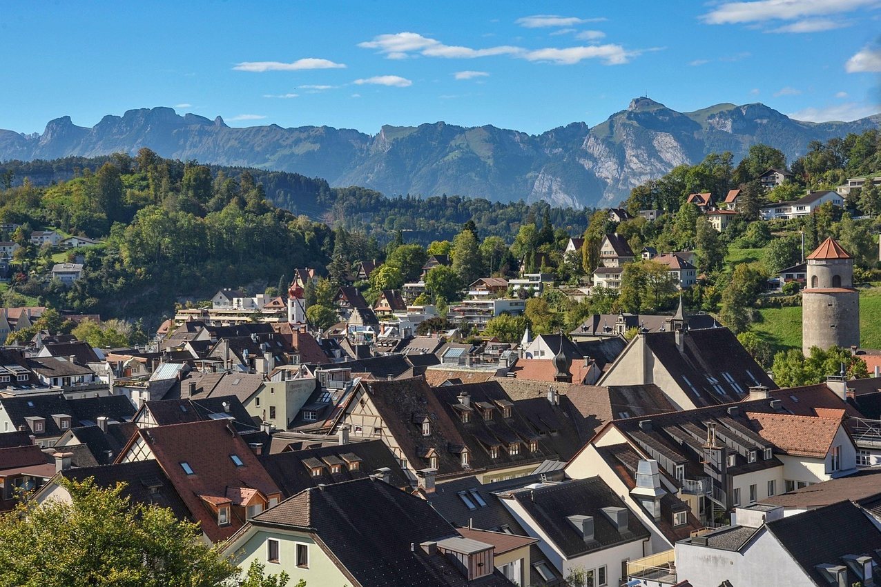 A picturesque European town with red-roofed houses nestled among green hills and towering mountains under a blue sky