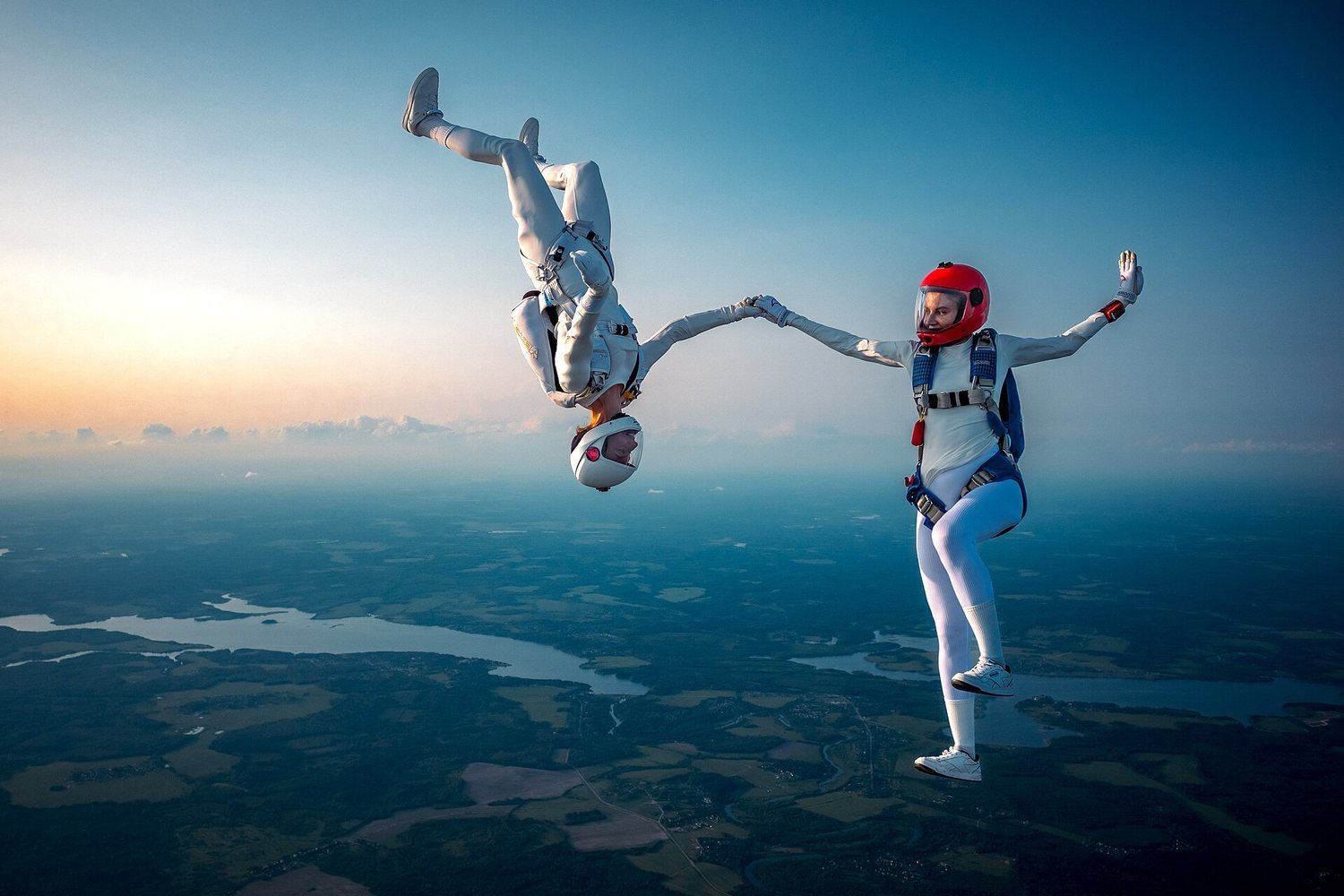 Two skydivers in mid-air, one upside down, holding hands against a sunset backdrop
