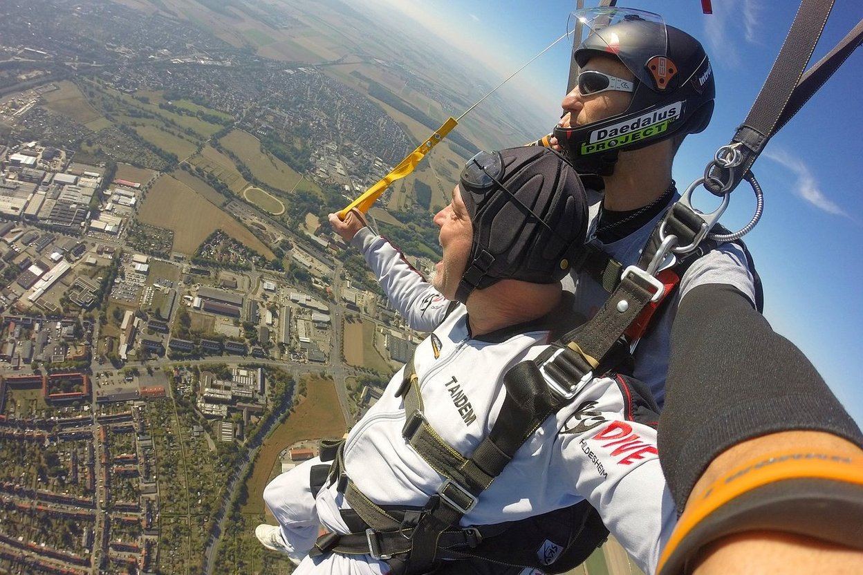 An older adult smiling in a tandem skydiving harness, with an instructor strapped to their back before boarding the aircraft.