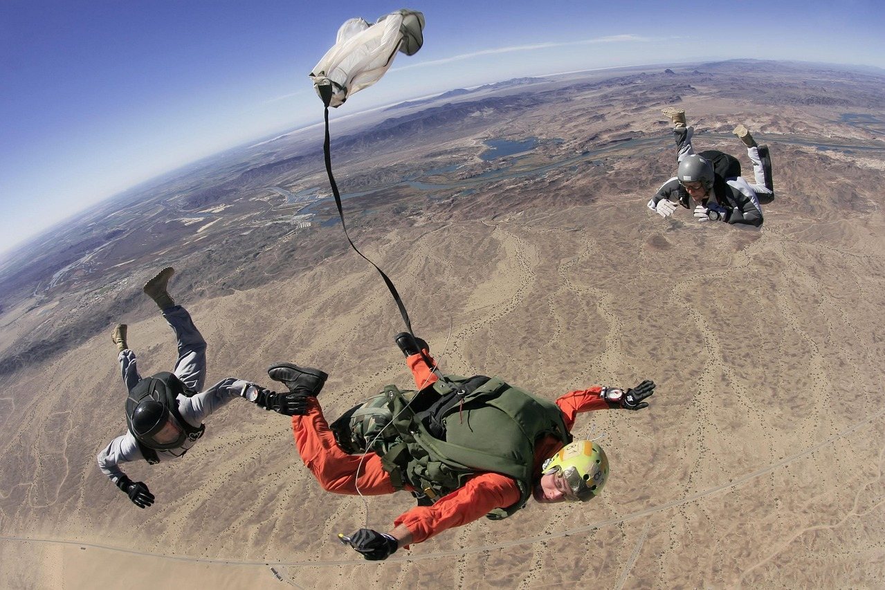 Three skydivers in freefall over a desert landscape with a parachute deployed