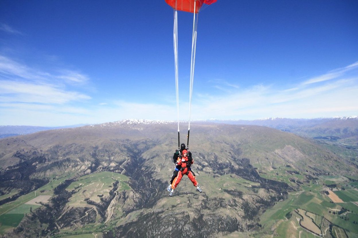Two skydivers under a red parachute over mountains on a clear day.