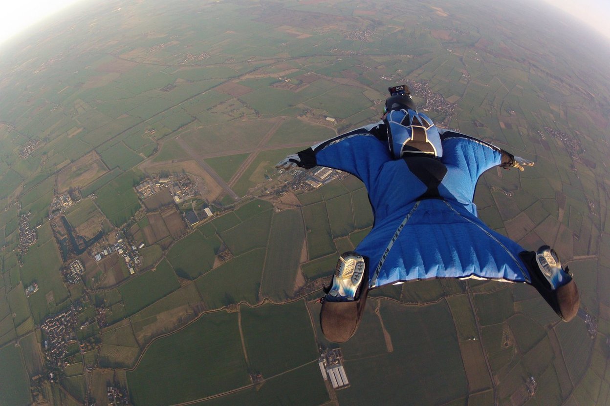 Wingsuit flyer glides over patchwork farmland below.
