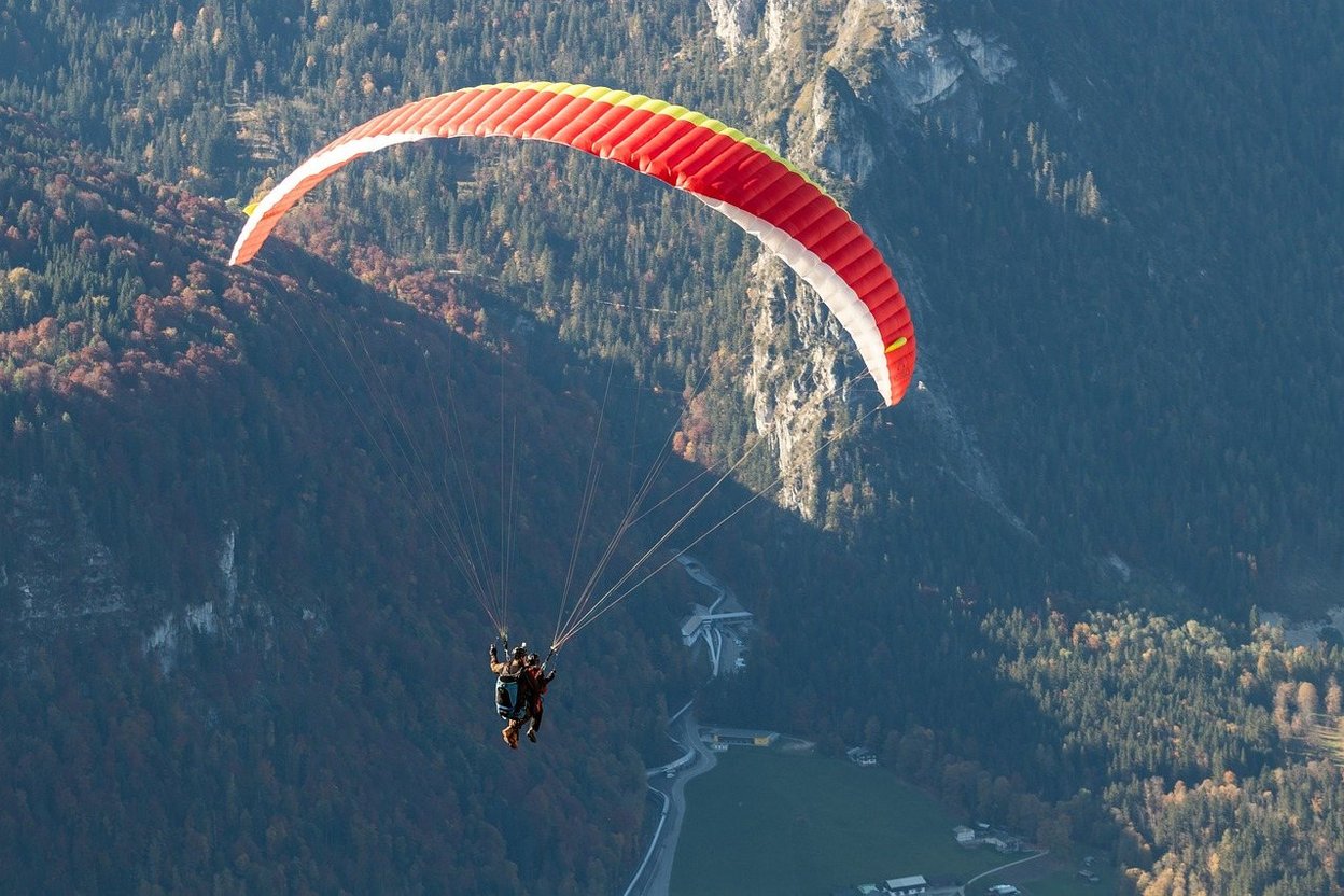 Red and white paraglider soars over forested mountains.