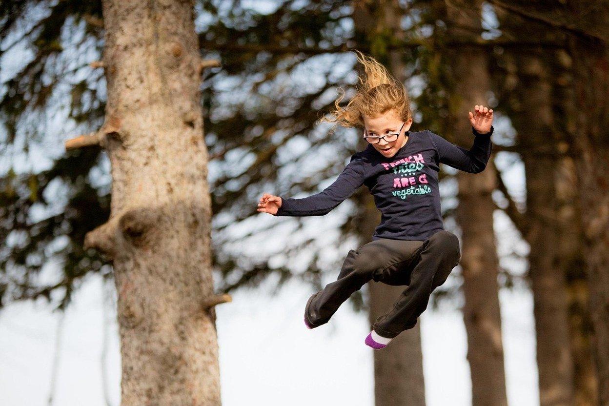 A girl leaps mid-air in a forest, hair flying, wearing glasses and a graphic shirt.